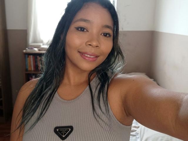 XinemaBonet met een medium bruine huid, lang zwart-groen haar, een klein zwart driehoekig logo op een grijze ribtop, lachend, roze lippen, met een arm uitgestrekt voor een selfie..