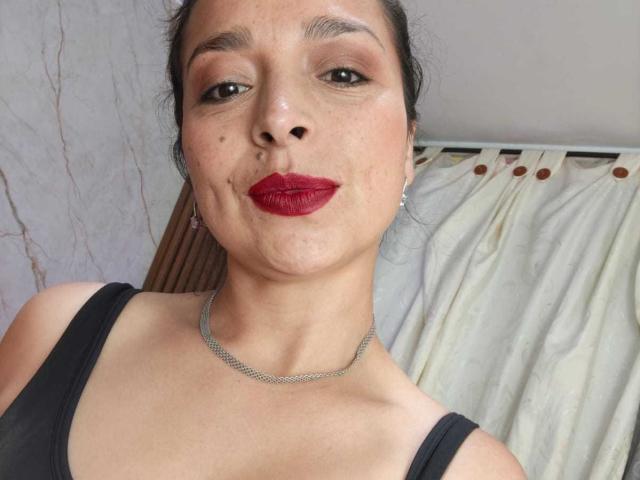 StangelarRivera has a pouty face, dark hair, red lipstick, small moles, a black tank top, and a silver necklace, standing in front of white curtains.