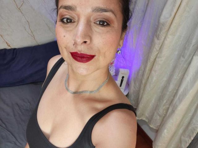 StangelarRivera with dark red lipstick, black tank top, silver necklace, and earrings, showing off her tits and slightly freckled face, with purple light in the background.