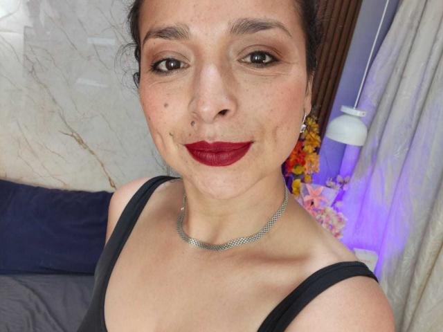 StangelarRivera has red lipstick, black tank top, silver necklace, and a nose ring. She's smiling with dark eyes and a slight double chin. Boobs are partially visible.