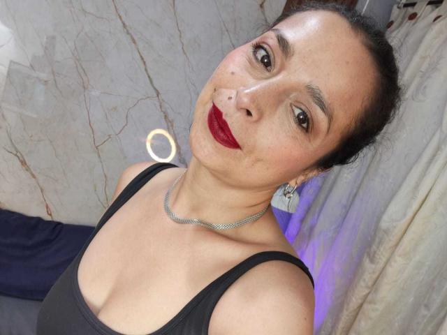 StangelarRivera with dark hair in a bun, red lipstick, black tank top, and silver necklace, showing cleavage, smiling at the camera, wearing earrings, in a well-lit room.
