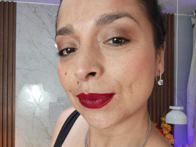 StangelarRivera has dark brown eyes, red lipstick, and a small mole on her left cheek. She wears black earrings and a silver necklace, with dark hair tied back.