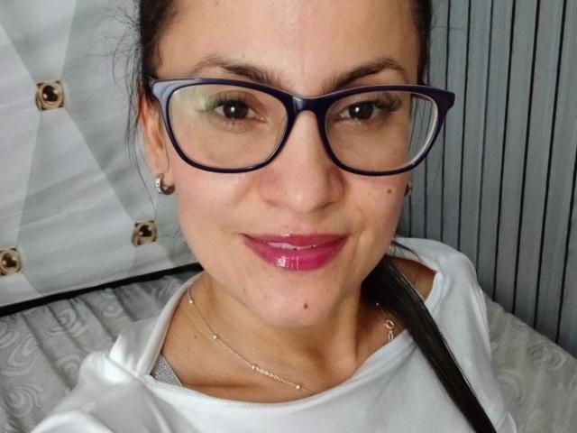 SolaraDubois with dark brown hair, blue glasses, pink glossy lips, white shirt, and necklace, smiling at the camera. Her tits are partially visible under the shirt.