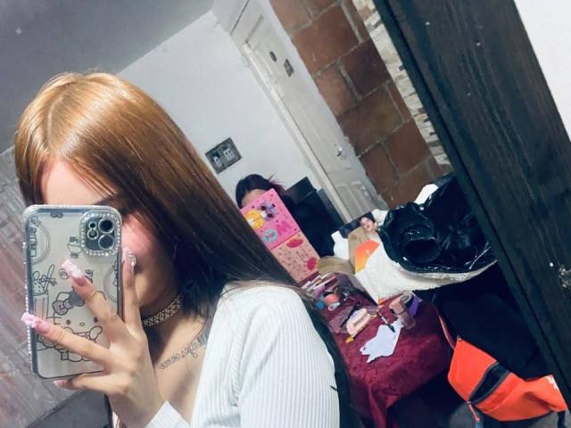 MadisonNoir with long brown hair, wearing a white top, holding a phone with a Hello Kitty case, taking a mirror selfie, showing off her tits and choker necklace.