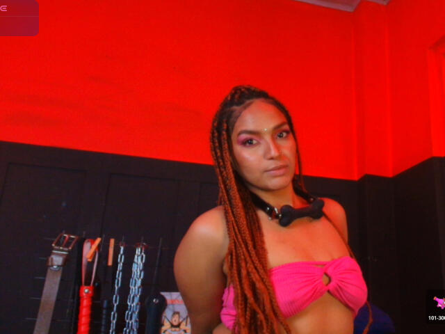 BrianaPratt with long orange braids, pink strapless top, black collar, and makeup, standing in red-lit room, looking serious, with BDSM gear in the background.