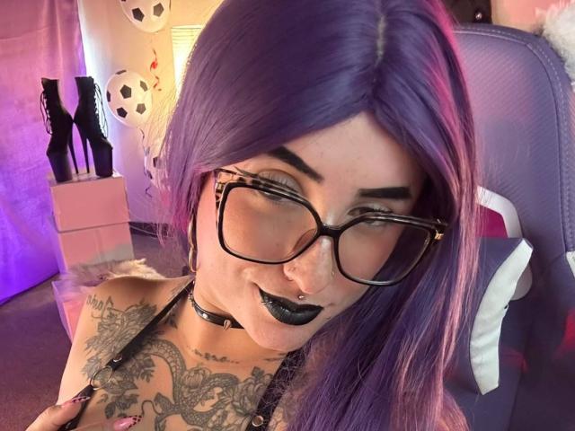 LilithDark69, avec des cheveux violets, du rouge à lèvres noir, des lunettes, un anneau au nez, un collier noir, une poitrine tatouée, tenant ses seins, assise sur une chaise violette, regardant la caméra..