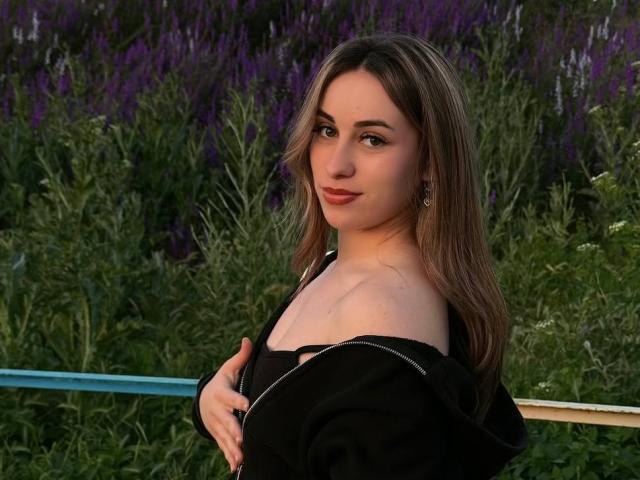 SunniTenen with long brown hair, off-shoulder black top, holding her tits, purple lavender in background, green foliage, subtle smile, earrings, soft evening light, casual outdoor setting.