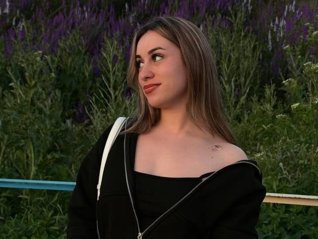 SunniTenen with long brown hair, black off-shoulder top, white strap, small tattoo on left shoulder, looking to the side with a slight smile, green and purple background.
