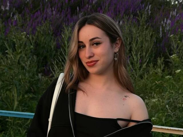 SunniTenen has light brown hair, a small tattoo on her right shoulder, and is wearing an off-shoulder black top, smiling with red lipstick, and a black jacket.