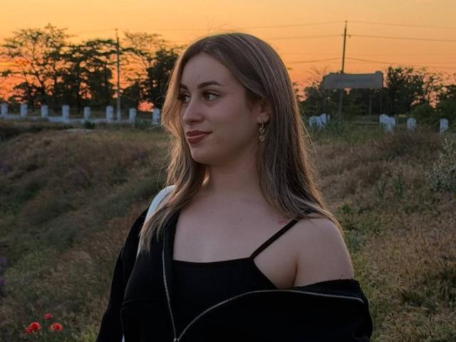 SunniTenen with medium tits, wearing a black off-shoulder top and black jacket, standing in a grassy field at sunset, looking to the side with a slight smile.
