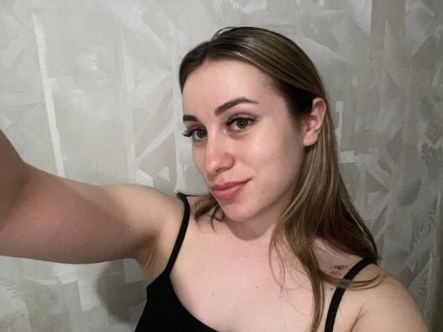 SunniTenen with light skin, brown hair, black tank top, small tattoo on right shoulder, smiling, holding arm out, eyeliner, subtle pink lipstick, medium-sized tits, gray textured wall.