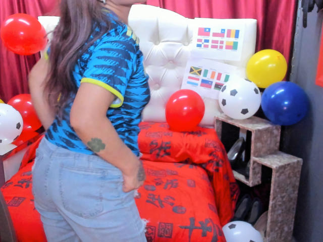 SofiaXDelRio vestindo uma camisa listrada azul e amarela, exibindo suas nádegas em shorts cinzas justos, com uma pequena tatuagem verde no braço esquerdo, cercada por balões..