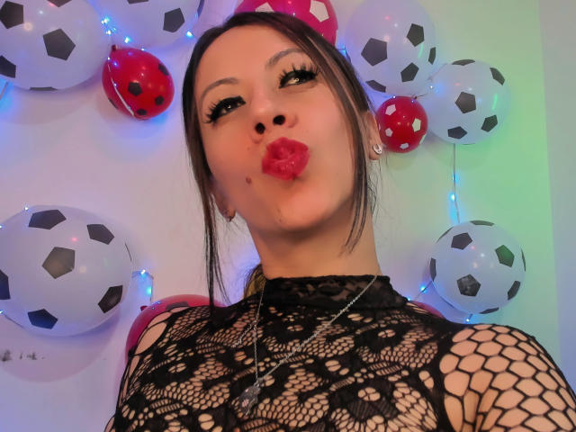 AmaraMirage, met volmaakte rode lippen, draagt een zwarte kanten top met netmotief en een ketting, en haar donkerbruine haar is in een paardenstaart gevlochten. Ze wordt omringd door ballonnen in de vorm van voetballen..