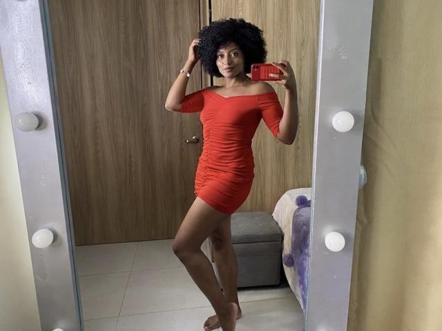RoldanStefy in a red off-shoulder dress, standing barefoot, holding a red phone, with curly black hair, looking in a mirror with bulb outlines.