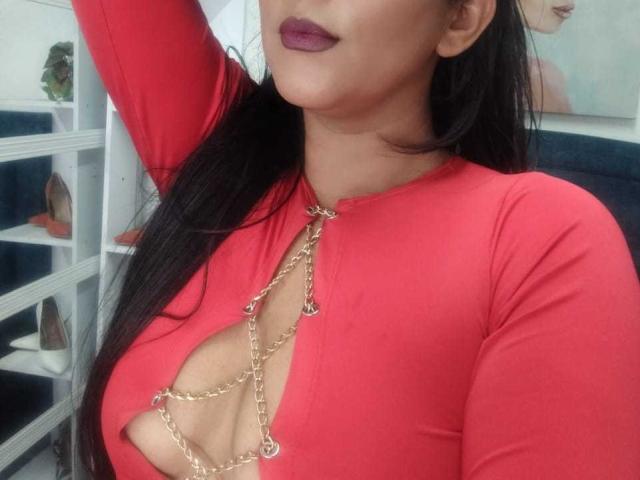 IsadoraVex mit dunklem Haar, einem roten Oberteil mit langen Ärmeln, einer Kette um den Hals, die das Dekolleté betont, lila Lippenstift, mit erhobenem Arm, wobei ihre Brüste sichtbar sind, in einem gut beleuchteten Innenraum..