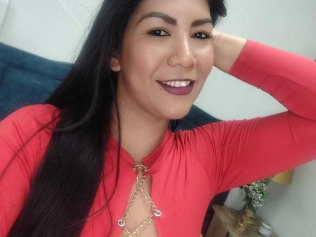 IsadoraVex, avec de longs cheveux noirs, un haut rouge, un collier en chaîne, souriant avec du rouge à lèvres violet, un anneau au nez, et une main derrière la tête, montrant ses seins à travers l'encolure du haut..