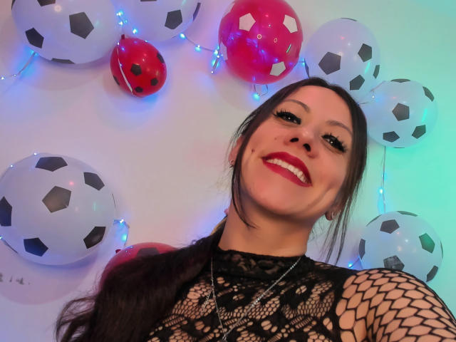 AmaraMirage has long dark hair, red lipstick, and is smiling at the camera. She's wearing a black lace top with fishnet sleeves, surrounded by soccer balls and red balloons.