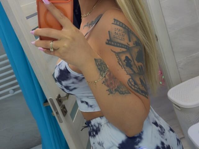 NicolSNaya with tattoos, holding orange phone, wearing tie-dye crop top and skirt, showing boobs, blonde hair, pink nails, gold chain, in bathroom mirror selfie.