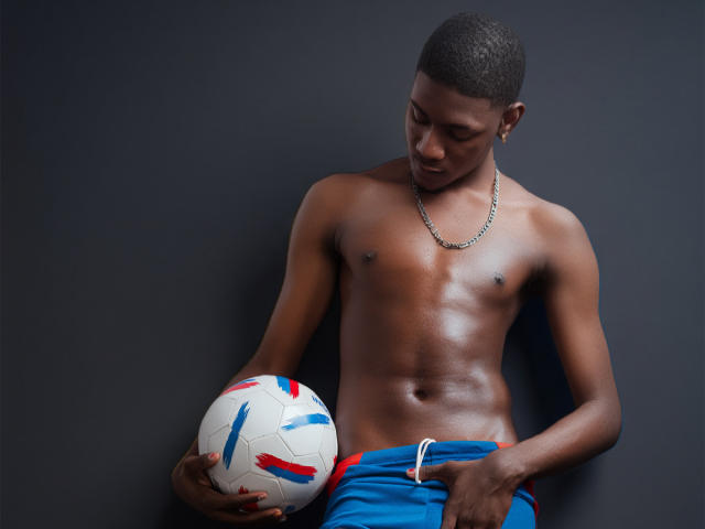 DrakeWit, shirtless with shiny dark skin, holding a soccer ball against his blue shorts. He's looking down, wearing a chain necklace, with a strong, muscular, sexy build.