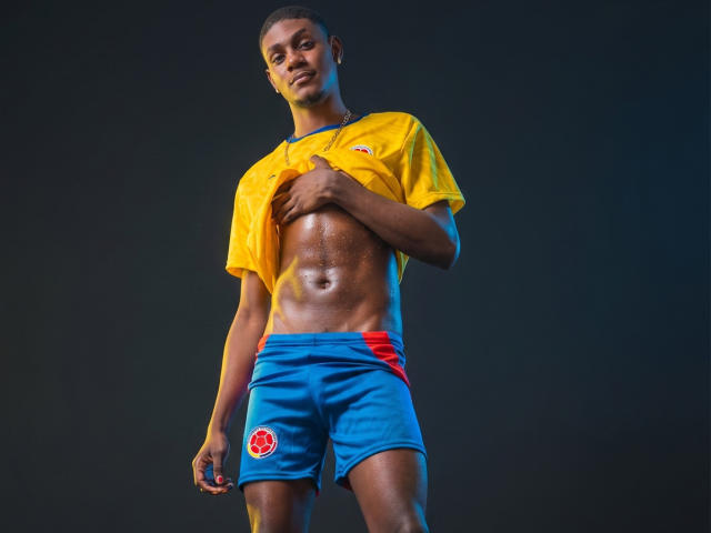 DrakeWit, un hombre negro musculoso, levanta su camiseta amarilla de fútbol, mostrando unos abdominales brillantes y definidos. Lleva pantalones cortos azules con un emblema rojo y está de pie con confianza..