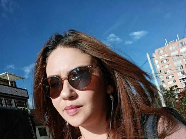 Theresa Larson, con cabello castaño, que lleva gafas de sol redondas y una blusa negra, tiene un piercing en la nariz y está haciendo pucheros, de pie al aire libre bajo un cielo azul brillante..