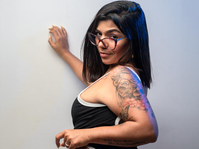 Valery Pierce, avec ses cheveux noirs, ses lunettes et son tatouage sur le bras droit, portant un débardeur noir, debout devant un mur blanc, regardant en arrière avec un léger sourire..
