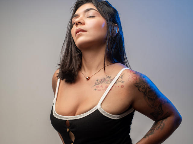 ValeryPierce with dark hair, eyes closed, wearing a black tank top with white trim, showing tattoos on her right shoulder and chest, small tits, and a necklace.
