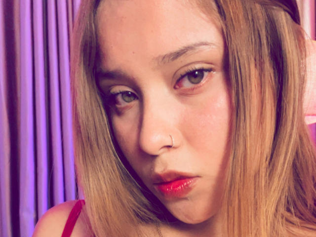 HeraJuno has pale skin, blonde hair, a nose ring, and red lipstick with a pouty expression, looking slightly to the side with blue eyes. Her tits are small.