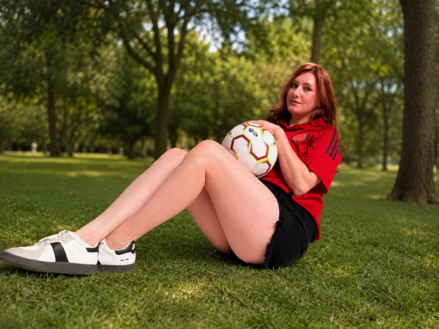 KatyaRogersX sits on grass with legs crossed, holding a soccer ball against her ass, wearing a red shirt, black shorts, and white sneakers with black stripes.