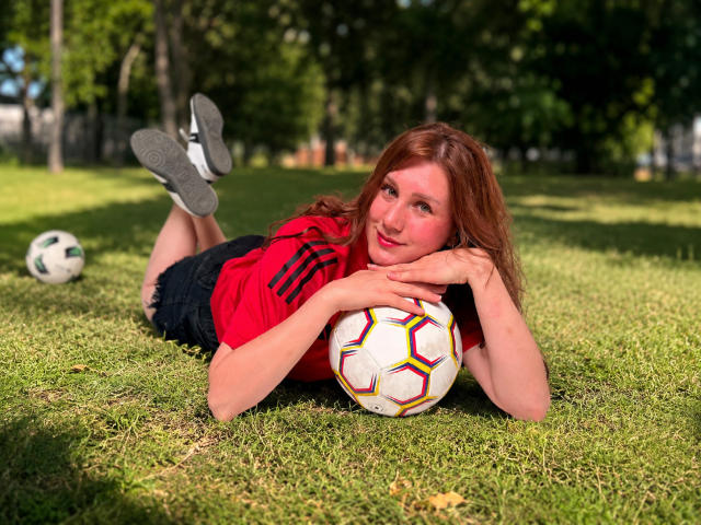 KatyaRogersX, red-haired girl, lying on grass, legs up, red shirt, black shorts, soccer ball under chin, smiling, hands on ball, white sneakers, green park, sunny day.