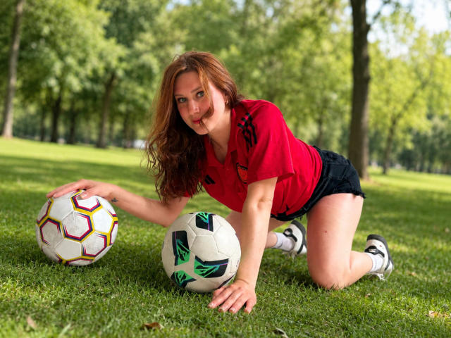 KatyaRogersX, with red shirt and black shorts, kneels on grass, touching two soccer balls, her brown hair loose, green eyes focused, and white socks with black shoes.