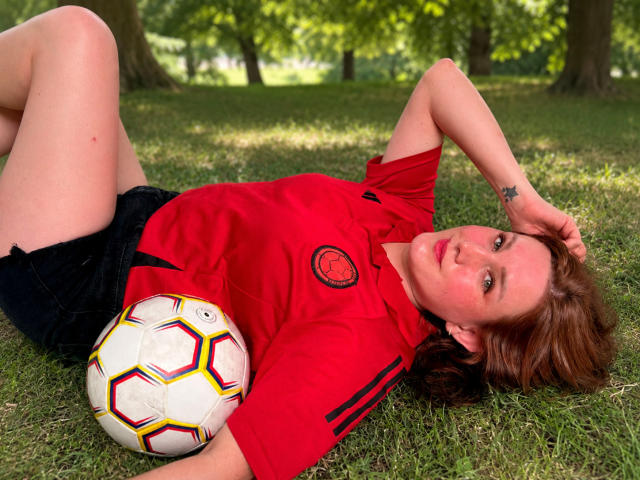 KatyaRogersX is lying on grass with a soccer ball, wearing a red soccer shirt and black shorts, her hand on her head, red hair, tattoo on her wrist.