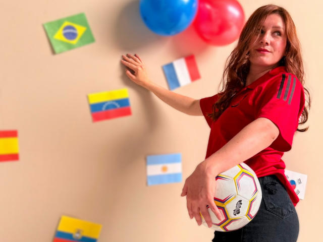 KatyaRogersX with red shirt, holding soccer ball, standing by wall with flags, red and blue balloons, long brown hair, looking up, hand on wall, jeans, slightly smiling.