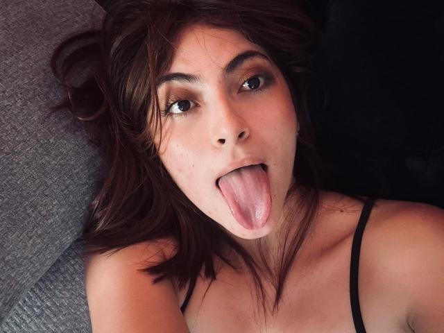 SpencerLe with brown hair, green eyes, and a tongue out, looking up, wearing a black spaghetti strap top, showing her bare shoulders and upper chest.