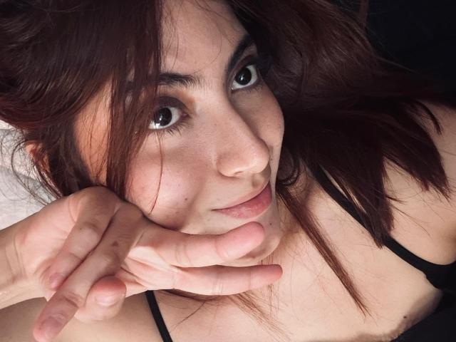 SpencerLe heeft bruin haar, grote bruine ogen en een schattige glimlach. Ze raakt haar kin aan met haar vingers, draagt een zwarte top met bandjes en laat haar borsten zien..
