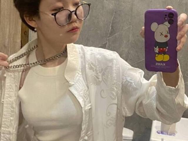 GladyBahde, portant des lunettes, une chemise blanche et une chaîne, tenant un téléphone violet avec un autocollant Mickey Mouse, se prend en photo devant un miroir et montre ses seins..