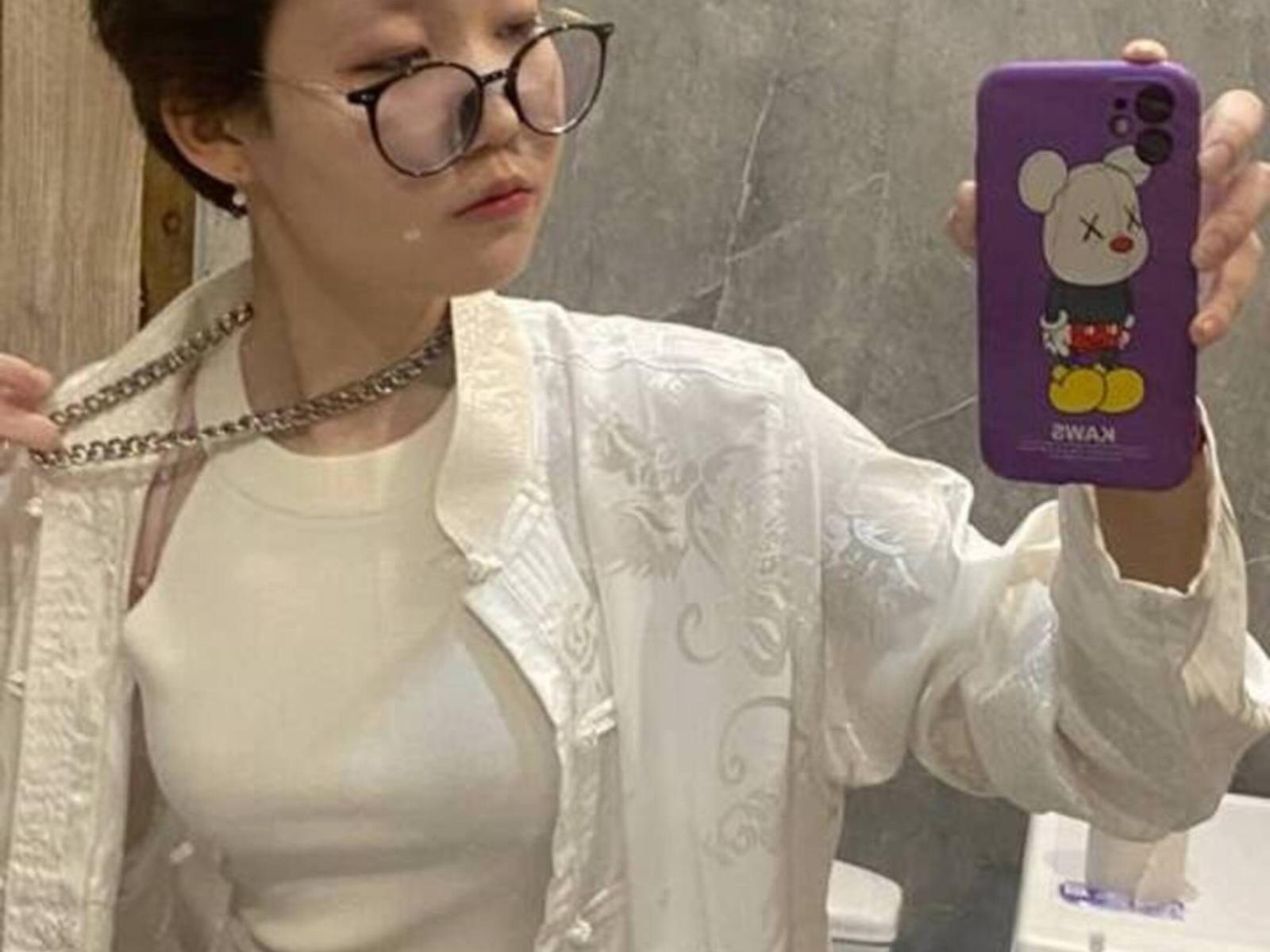 GladyBahde, usando óculos, camisa branca e um colar, segurando um telefone roxo com um adesivo do Mickey Mouse, mostrando seus seios em uma selfie no espelho..