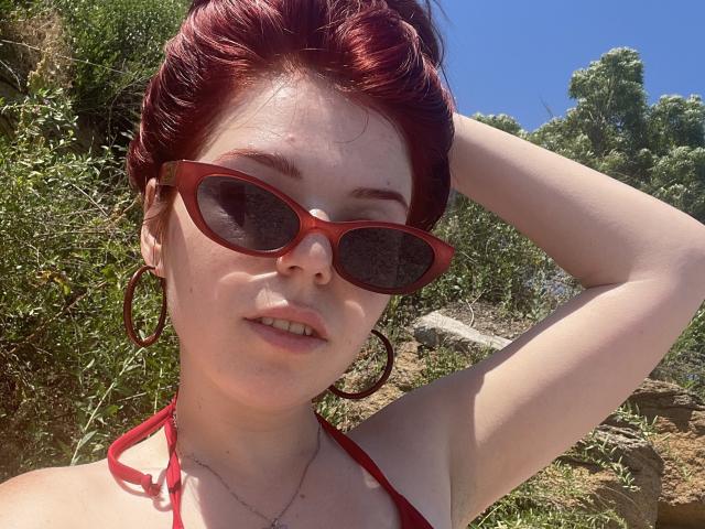 AlexXas, con i capelli rossi, gli occhiali da sole rossi e il top a fascia rosso, che mostra il seno, ha una mano tra i capelli, indossa grandi orecchini a cerchio, in un ambiente esterno soleggiato..