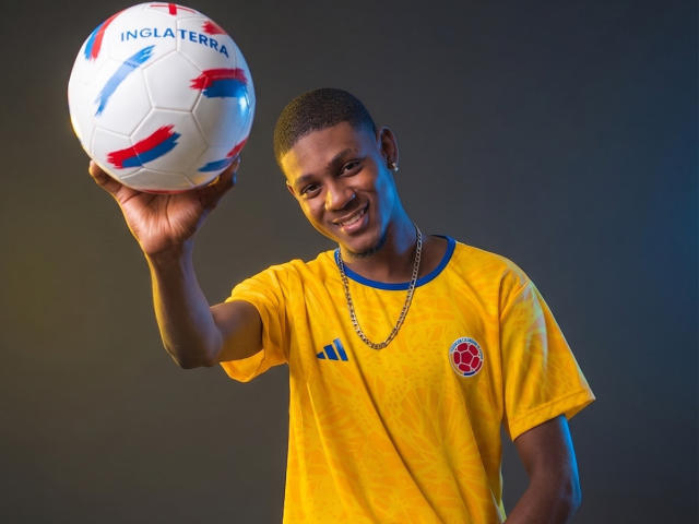DrakeWit with short black hair, smiling, holding a white soccer ball with red and blue stripes, wearing a yellow soccer jersey, and a chain necklace.
