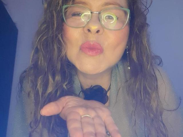 MiaAmoretti met krullend bruin haar, groene bril en een roze mond, die een kus maakt, draagt een grijze top en houdt haar hand met een ring omhoog, met daarin een zwarte roos..