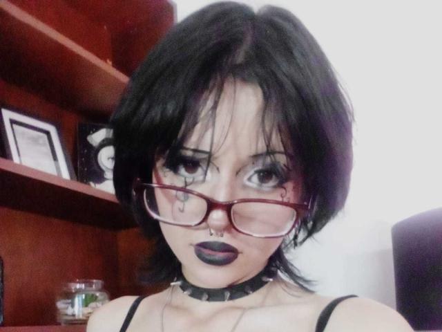 ViolettClay has black hair, red glasses, black lipstick, a nose ring, and a spiked choker. Her tits are small, and she has a tattoo under her right eye.