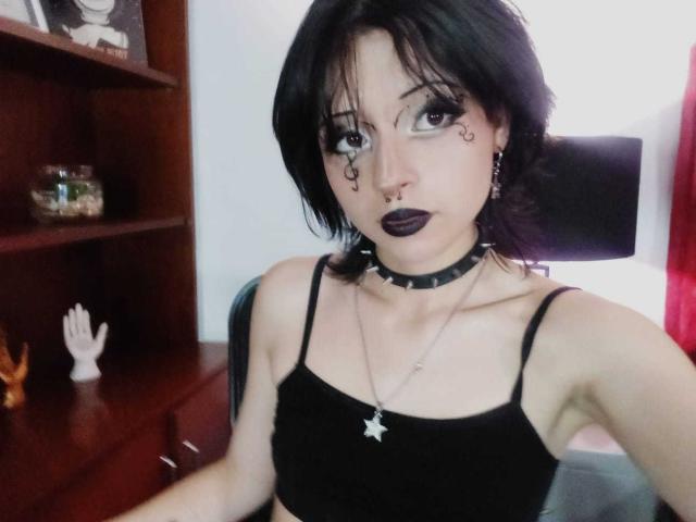 ViolettClay with black hair, goth makeup, black lipstick, black choker, star necklace, black bra, small tits, sitting, looking up, wooden shelf with fishbowl in background.