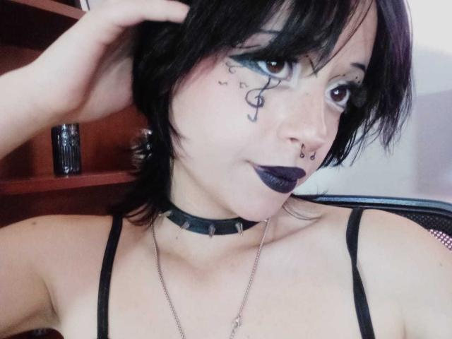ViolettClay with black hair, dark eye makeup, purple lipstick, black choker, nose ring, and tattoos, wearing a black strap top, sitting in a chair.