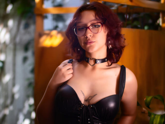 JinXkm met rood krullend haar, een bril, een zwarte leren bustier, een choker en een tatoeage boven de borsten, houdt een band vast en kijkt naar de camera..