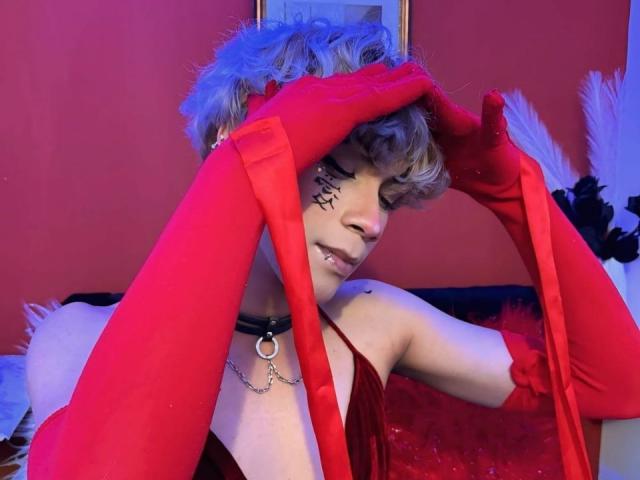 SoyCooper, con cabello corto, rizado y azul-grisáceo, vistiendo guantes rojos, un vestido rojo, un collar negro con un anillo, y tatuajes faciales negros, cubriendo los ojos con las manos, en una habitación roja..