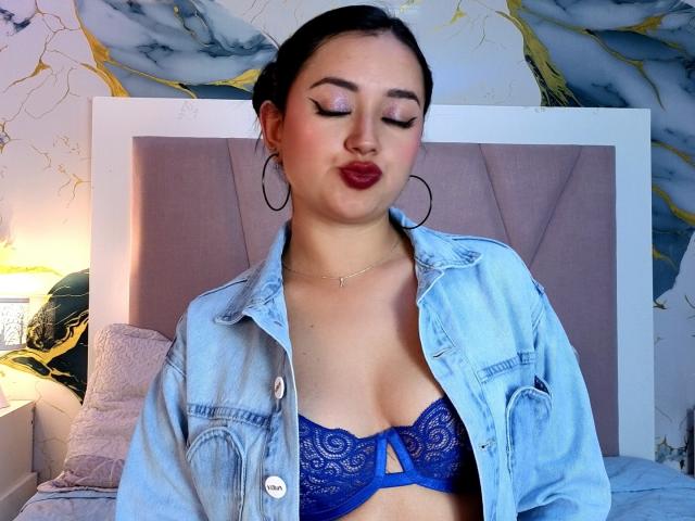 MiiaRousse, com os olhos fechados, vestindo um sutiã de renda azul, jaqueta jeans aberta e batom vermelho, está em pé em um quarto com cabeceira cinza e papel de parede de mármore amarelo..
