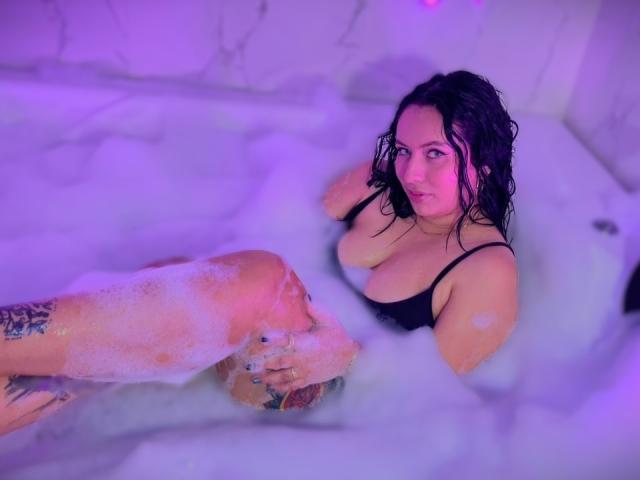 Amy Ross, con el cabello negro y mojado, en una bañera llena de burbujas, vestida con un top negro de tirantes, mirando hacia arriba, con los senos parcialmente visibles, un brazo tatuado apoyado en su muslo..