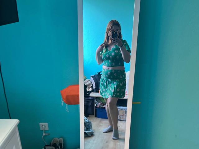 Jamilia, vestita con un abito verde a fiori con maniche corte e una gonna, tiene un telefono davanti a uno specchio, mostrando il seno e i glutei. Indossa calze bianche..