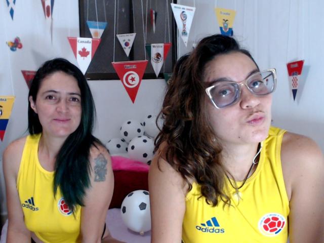 LunaVenus, avec des cheveux noirs aux pointes vertes, de petites seins et un tatouage, portant un t-shirt Adidas jaune, est assise à côté de LunaVenus, avec des cheveux bruns bouclés, des lunettes et une moue, également vêtue d'un t-shirt jaune..
