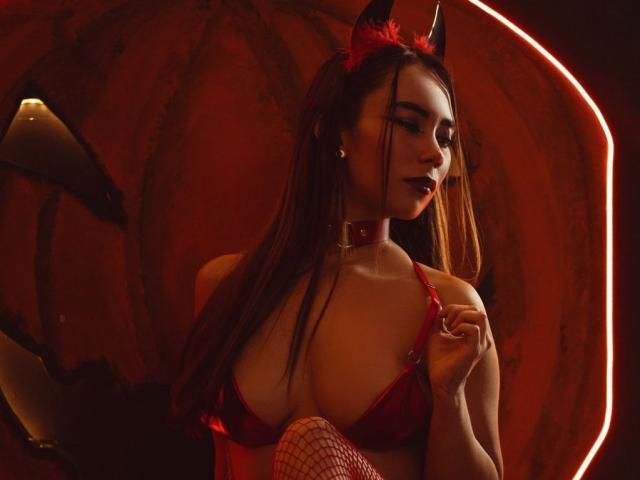 SophiaSmiht com chifres vermelhos de diabo, sutiã vermelho, meias de rede, e colar, segurando a alça do sutiã, mostrando os seios, cabelo castanho longo, batom escuro, fundo vermelho brilhante..